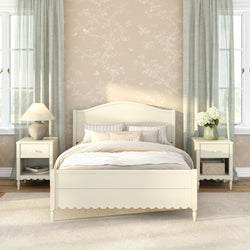Cozy cream farmhouse bedroom with wooden bed, matching nightstands, beige floral wallpaper, sheer green curtains, table lamp and vase of white hydrangeas