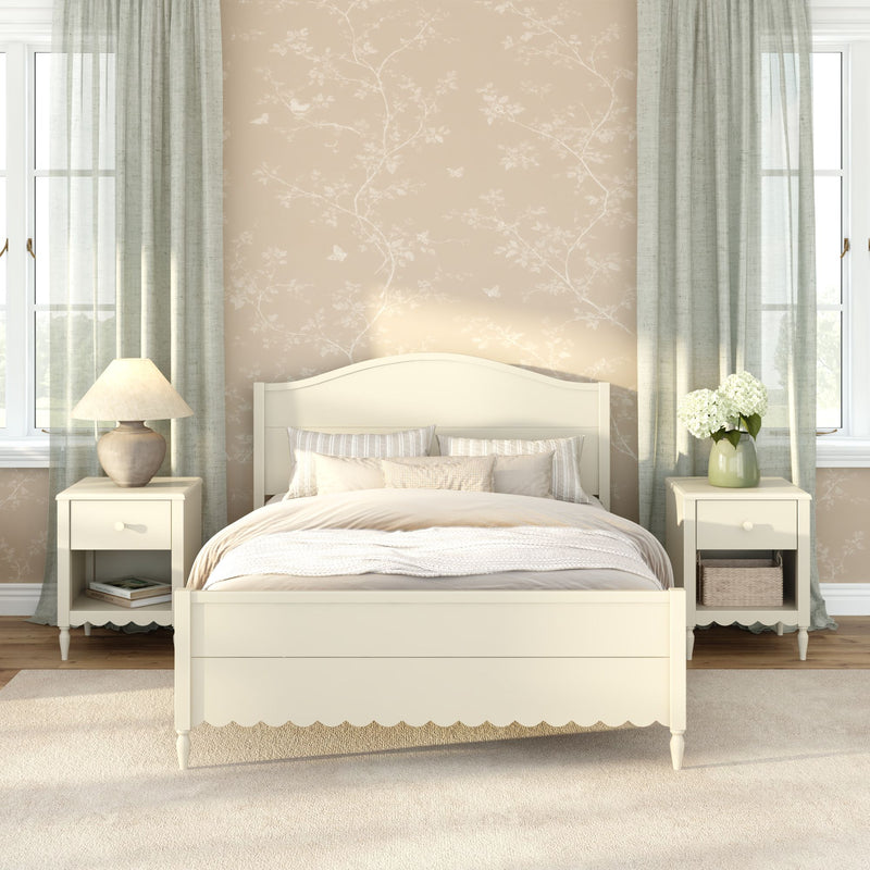 Cozy cream farmhouse bedroom with wooden bed, matching nightstands, beige floral wallpaper, sheer green curtains, table lamp and vase of white hydrangeas