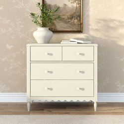 Cream wooden chest of drawers with scalloped trim and round knobs, styled with a ceramic vase of greenery and books on a hardwood floor against neutral floral wallpaper — modern farmhouse bedroom dresser decor
