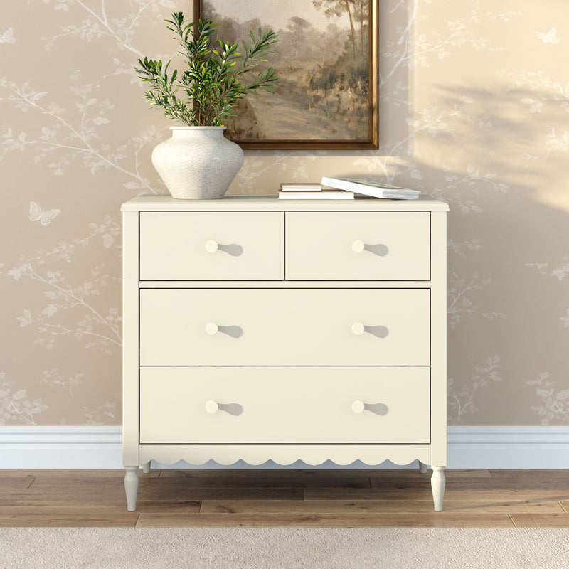 Cream wooden chest of drawers with scalloped trim and round knobs, styled with a ceramic vase of greenery and books on a hardwood floor against neutral floral wallpaper — modern farmhouse bedroom dresser decor