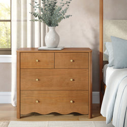 Wooden bedroom dresser nightstand with brass knobs topped by a ceramic vase of eucalyptus, light blue pillows and neutral curtains