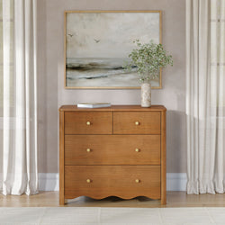 Natural wood dresser with brass knobs beneath a framed coastal abstract painting with flying birds, marble vase of green stems and soft linen curtains in a neutral minimalist bedroom or entryway decor.