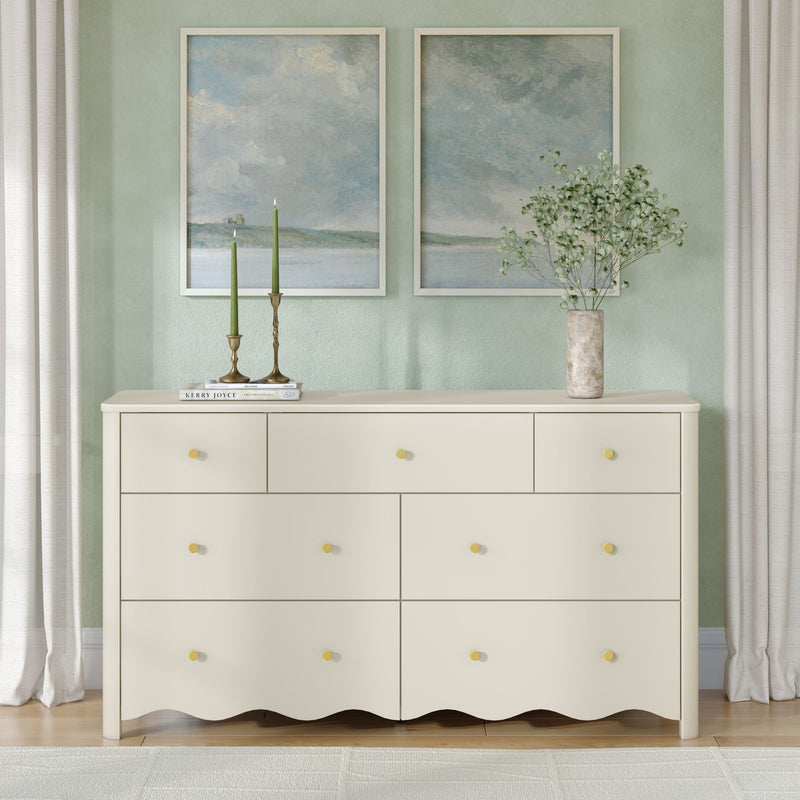 Cream wooden dresser with yellow knobs styled with brass candlesticks and green taper candles, marble vase of delicate greenery, and twin framed coastal art prints on a pastel green wall with neutral curtains