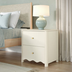Close-up of a white bedside nightstand with gold knobs, pale blue ceramic table lamp and soft blue pillows against a beige upholstered headboard and curtains, modern coastal bedroom decor