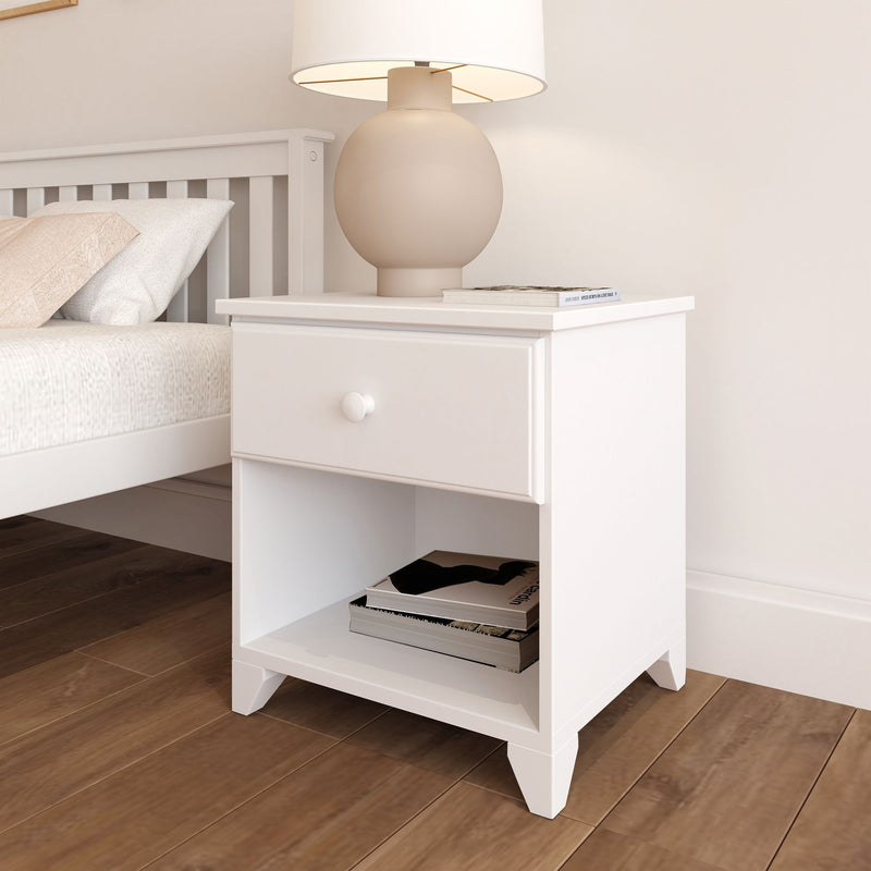 White wooden nightstand with a drawer and shelf, featuring a round lamp on top next to a bed with white linens and pillows.
