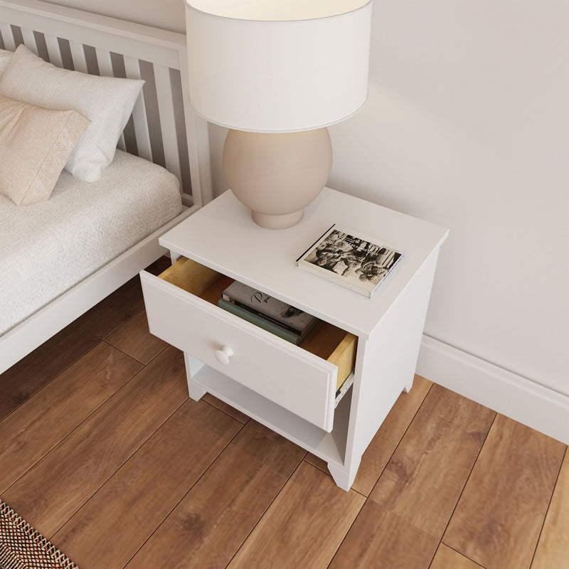 White nightstand with open drawer, modern lamp, and book on wooden floor beside bed.