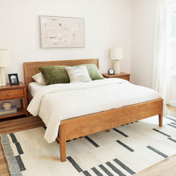 Modern minimalist bedroom with wooden bed frame, cream bedding, green velvet pillows, matching nightstands, table lamps, abstract wall art and geometric area rug