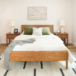 Minimalist modern bedroom with wooden bed frame, layered white bedding, green velvet pillows, matching wood nightstands, ceramic lamps and abstract wall art