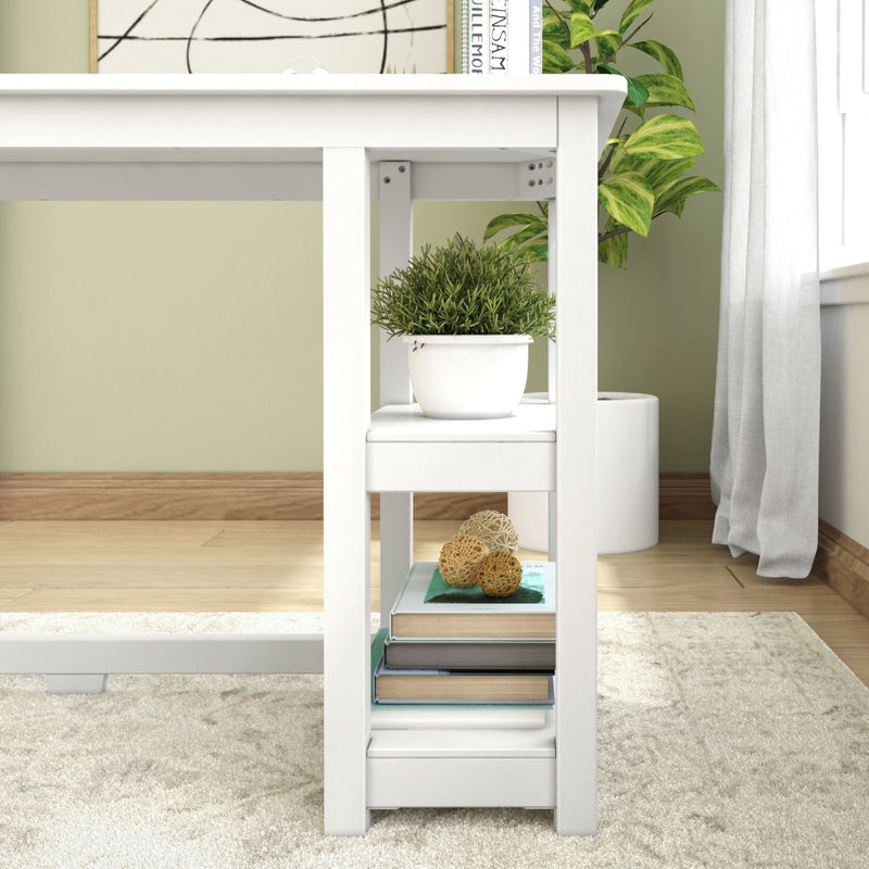 White wooden desk with built-in lower shelf holding a stack of books, decorative rattan balls and a potted green plant in a white pot in a bright modern home office with light hardwood floors and beige rug
