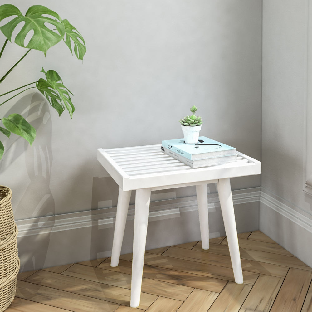White slatted side table with potted succulent and books, next to green plant in woven basket, on wooden floor.