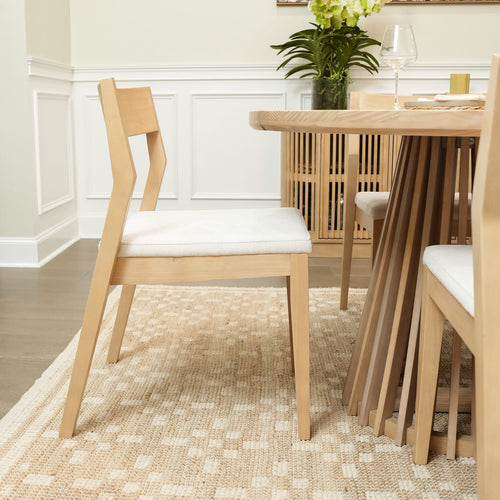 Modern light wood dining chair and round wooden dining table on beige woven rug in bright dining room with white wainscoting and potted plant.