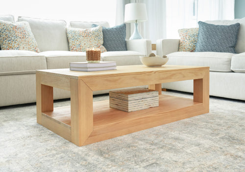 Light wood rectangular coffee table with lower shelf styled with stacked books, a wicker candle and decorative bowl in a modern neutral living room with beige sofa and patterned throw pillows.