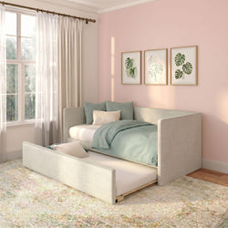 Pastel pink bedroom with upholstered daybed and pull-out trundle, mint-green bedding, sheer curtains, and framed monstera botanical prints in soft natural light.