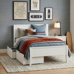 Modern bedroom with white platform bed and under-bed storage drawers, blue quilt and rust knit throw, decorative pillows, framed gallery wall, bedside lamp and skateboard accent