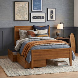 Wooden storage bed with blue and tan layered bedding, textured rug, nightstand lamp and gallery wall featuring skateboard art and vintage map