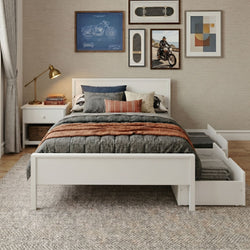Modern neutral bedroom with white platform bed, layered gray and terracotta bedding, textured pillows, brass bedside lamp, motorcycle blueprint and skateboard wall art above bed, under-bed storage drawer