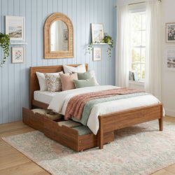 Wooden storage bed with pull-out drawers in a cozy coastal bedroom featuring pastel floral bedding, blue shiplap wall, rattan arched mirror, framed botanical art, trailing houseplants and sheer white curtains.