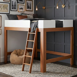 Modern wooden loft bed with ladder, charcoal bedding and leather accent pillow against a dark gray paneled wall with brass sconces, gallery art and corduroy bean bag