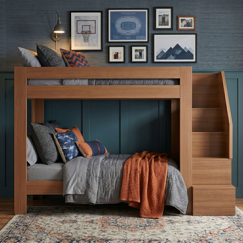 Wooden bunk bed with built‑in stairs against navy blue paneled accent wall, gray quilted bedding, patterned pillows and orange knit throw, gallery wall of framed art, modern kids' bedroom decor