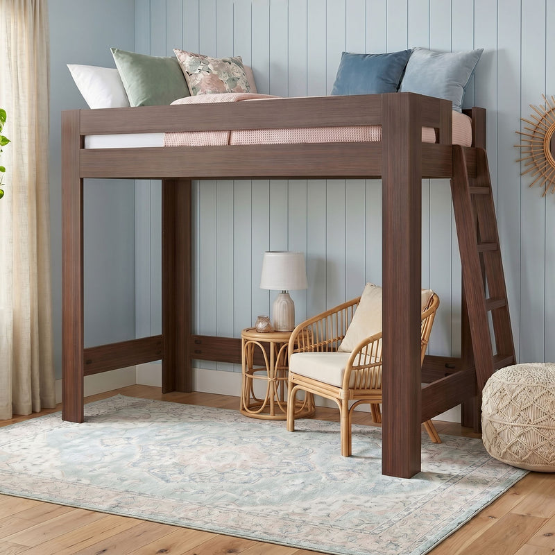 Walnut wooden loft bed with ladder, pastel pillows and pink knit blanket above a rattan seating nook and lamp against a light blue shiplap wall on a patterned area rug — modern kids' bedroom design