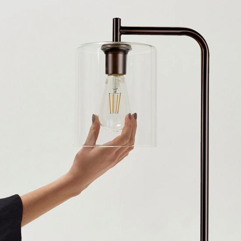 Hand adjusting clear glass cylinder shade with LED filament bulb on bronze metal floor lamp, modern industrial lighting fixture